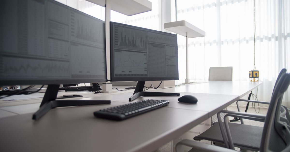 An office desk with dual monitors showing data, a keyboard and ergonomic chairs
