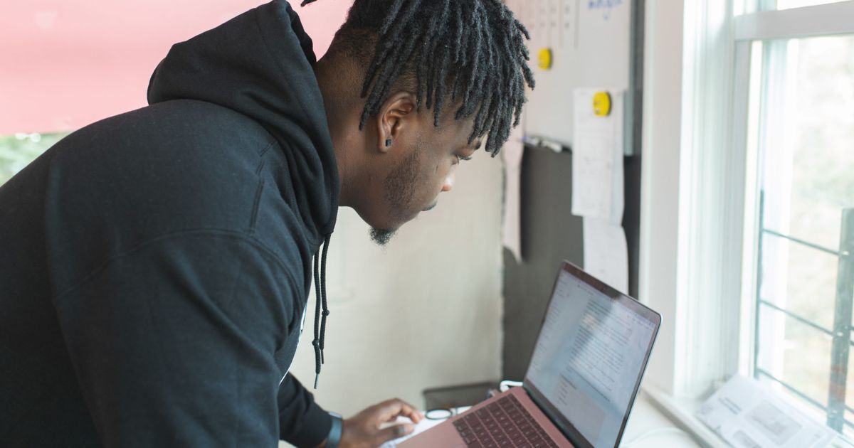 A man in a dark hoodie leaning over a laptop in a bright home office