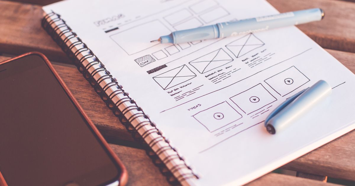 A notebook with hand-drawn website wireframes beside a smartphone on a wooden desk